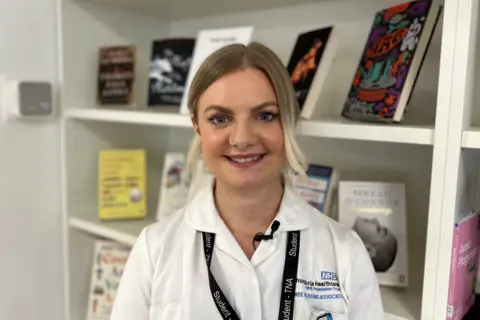 Laura Hurst is wearing a white medical jacket with a lanyard around her neck indicating that she is a student. She is pictured in a library. She has long blonde hair that is tied in a ponytail and is smiling. 