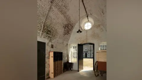 Historic England The inside of a room, with an arched brick roof that has been painted white. There are a number of stains on the brickwork suggesting damp. Inside the room is what appears to be a horse cart and an empty bookcase. Through an open door you can see the inner courtyard to the fort. 