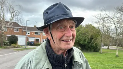 Kimberly Piper/BBC 91-year old Glyn Parry is stood outside smiling at the camera. He is wearing a hat with a piece of shoelace tied underneath his chin to keep his hat on. He is standing in front of a number of houses, by a green area with trees.