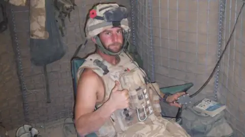 Major John Whiteman in uniform in a military shelter with a helmet on and sitting on a green chair.