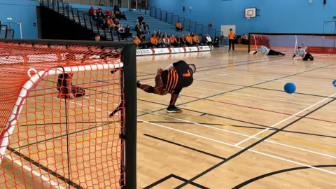 A goalball game in progress, a man in a black and orange strip throws a blue ball across a sports hall. 