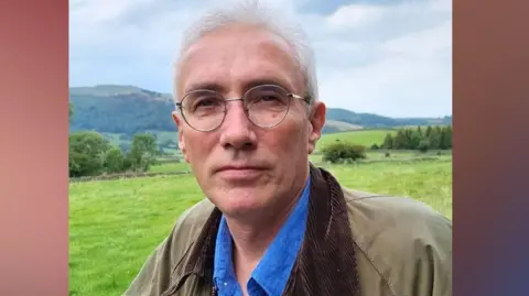 Alan Cleaver Alan Cleaver, with short white hair, and glasses, wearing a brown jacket, looks at the camera. Behind him are green fields and hills. 