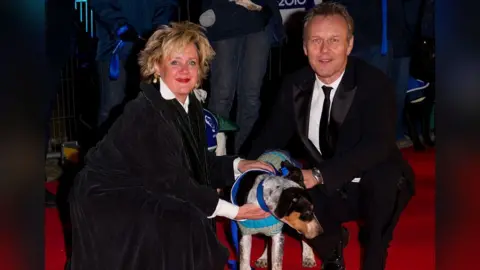 Getty Images Anthony Head and Sarah Fisher attending Collars and Coats Gala Ball to mark the 150th Anniversary of Battersea Dogs and Cats home in 2010. Sarah is wearing a dark velvet style cape and a white blouse and Anthony is wearing a black suit, white shirt and tie. They have a white dog with brown and black markings, wearing a dog jacket between them. The dog is looking down to the floor and Sarah and Anthony are looking at the camera smiling. 