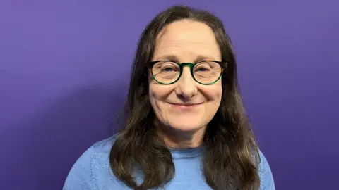 A woman stood in front of a purple wall. She has long brown hair and glasses and is wearing a pale blue top