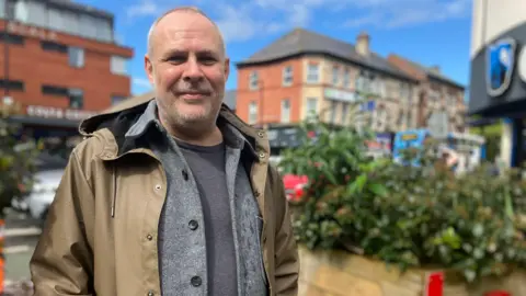 Prof Steve Millington stands by some shrubs on a sunny day on Withington High Street