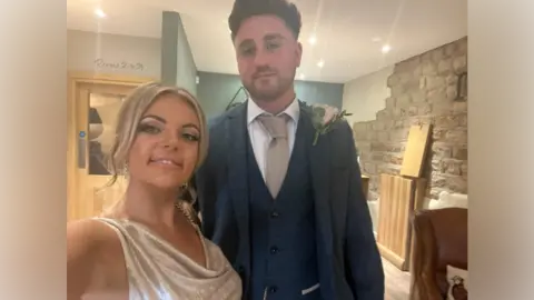 A young woman with blonde hair and a glittery dress, stands next to a young man in a navy suit inside a pub