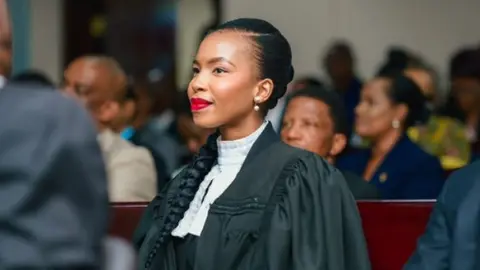 Lesego Chombo/Instagram Lesego Chombo in a black legal gown and white collar. Wearing red lipstick, she is seated on a bench with others in a courtroom and is looking to the side - not at the camera.