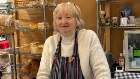 Jenny has short white blonde hair with glasses on her head. She wears a cream jumper and an apron with black, blue and pink strikes with orange flowers on it. Behind her are metal shelves with various golden loaves of bread on them