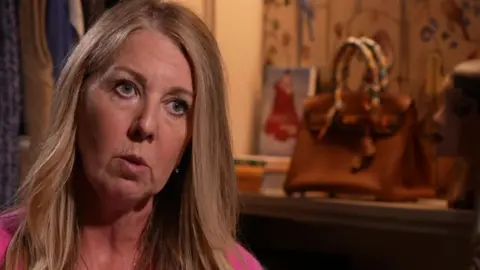 Christine Colbert who has long dark blond hair and wears a cerise pink cardigan and top is sitting in a shop with a rail of clothes behind her and a designer beige handbag with multi-coloured straps on a shelf in the background.