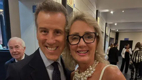 Warren Bullock Anton Du Beke and Jane Bullock, a man in a black suit, grey shirt and black spotted tie is smiling next to a blonde woman with black rimmed glasses and a large pearl necklace