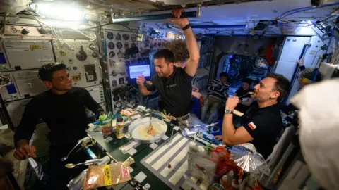 Axiom Space Indian astronaut Shubhanshu Shukla with his Ax-4 colleagues at ISS engaged in a conversation with food packets and plates on the table in front of them.