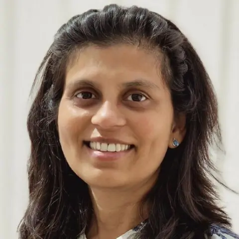 Priya Kapadia, con el pelo largo y negro recogido hacia atrás con una diadema, sonríe a la cámara.