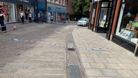 Blue police tap attached to cones and a building has cordoned off a section of a city street in Norwich, outside a Waterstone's store, which has black and white branding, with books in the windows on display. A police car can be seen in the background next to the cordon, as people walk past it. 