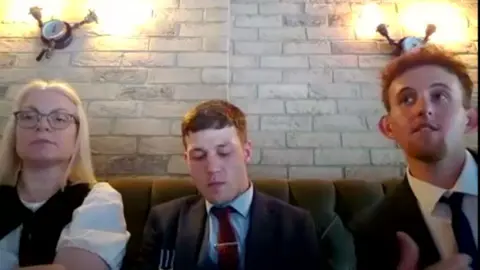 Caralyn Jones (L), Luke Wood and Kevin Macdonald are sitting on a sofa in a restaurant wearing smart clothing. 