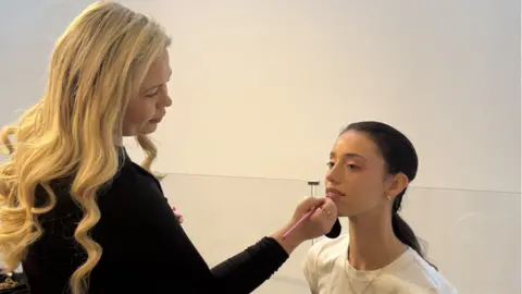 Georgia Sullivan Georgia applying lipliner onto a woman's lips. Georgia, on the left, wears a long sleeved black top and has long blonde hair. The model wears a white t-shirt, silver necklace and has black hair in a ponytail. 
