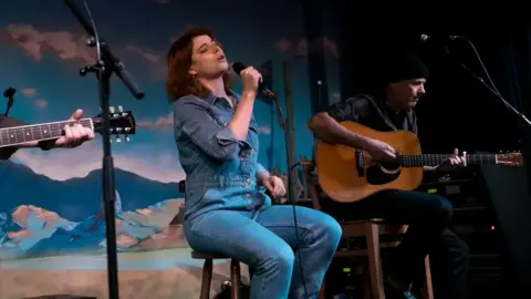 Eoin Carey Jessie Buckley, who has brown shoulder-length hair and is wearing denim jumpsuit, sits on a stool between two guitarists and holds a microphone as she performs at the premiere of Wild Rose in Glasgow. 