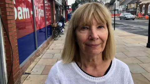 John Devine/BBC Julie Gargiulo is stood outside the Tesco Express on March High Street wearing a white jumper with a black top underneath. She has straight shoulder length blonde hair and a fringe. Behind her right shoulder you can see construction workers.  