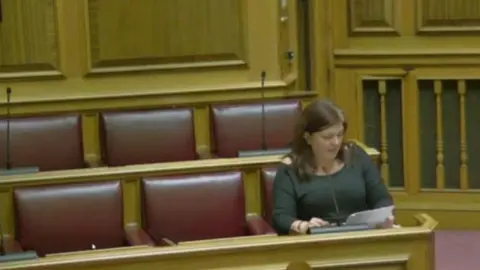 Dorset Council Unison rep Becky Brookman sitting alone in part of the council chamber. She is sitting at the end of a bench of red leather seats. She is reading from a piece of paper. She has long dark hair and is wearing a dark long-sleeved top.