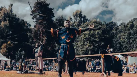 RJJonesPhotography Henry Browne is dressed in medieval knight armour with navy overalls with orange crown logos on it. His arms are stretched out either side of him and he is holding a long sword in one of his hands. Behind him are other people dressed in medieval clothes as well as two black horses and a crowd of people sitting on the grass behind some bunting and fencing.