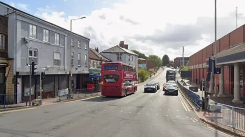 Google The road where Patricia Lashley was last seen, taken from a junction. Buildings are on both sides of the road, with a double-decker bus travelling away on the left and other vehicles are coming towards the camera on the right.