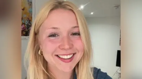 Aneesa Aneesa is a young woman with long blonde hair worn down. She smiles at the camera and wears a denim top and gold necklace and earrings