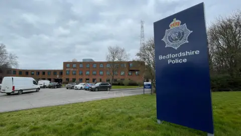 The headquarters of Bedfordshire Police in Kempston. There is a large, blue sign in the foreground which reads "Bedfordshire Police". There is a red brick building in the background with a telegraph mast next to it. There are cars and vans parked outside.