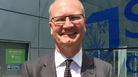 Matthew Hicks is standing in front of a glass building wearing a grey suit, white shirt and black and yellow tie. Hicks is wearing glasses and is smiling at the camera