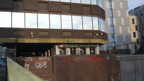 The Hustler Pool and Snooker club is part of a tall glass and red-brick building. The walls surrounding are dotted with graffiti. The signs on the venue's exterior have colourful snooker balls on them.