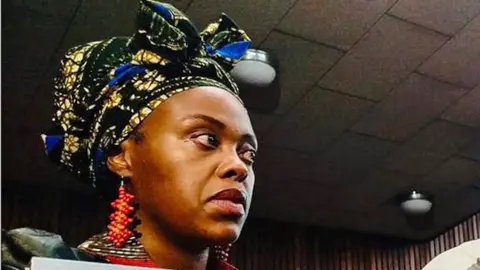 NPA Nonkululeko Patricia Mantula, wearing a headwrap, large beaded orange, red earrings and a leather jacket look on in court.