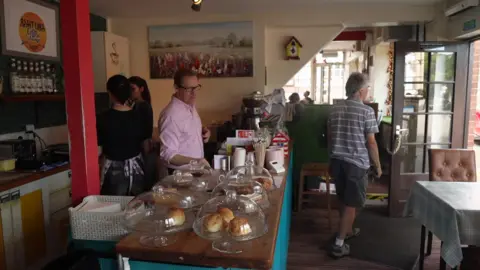 Interior of a café with a wooden counter displaying cakes and pastries under glass domes. Several customers and staff are visible around the counter and seating area. The café has colourful décor, framed artwork on the walls, and daylight entering through the front doorway and windows.
