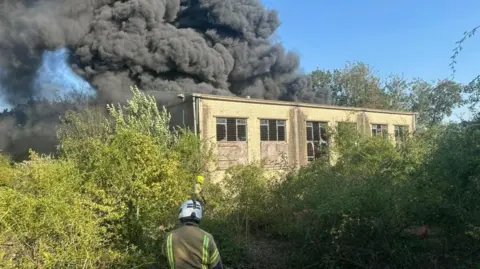 Essex County Fire and Rescue Service A fire officer in protective clothing and helmet stands in front of a pale-coloured one storey building, with their back to the camera. Smoke billows from the back of the unit. Shrubbery surrounds the site.