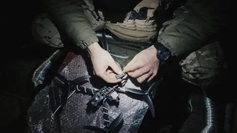 Volodymyr Rakov A soldier fixing or setting up a black drone in the dark. Only his hands and knees are visible, lit up by what seems to be a head torch shining down from above.