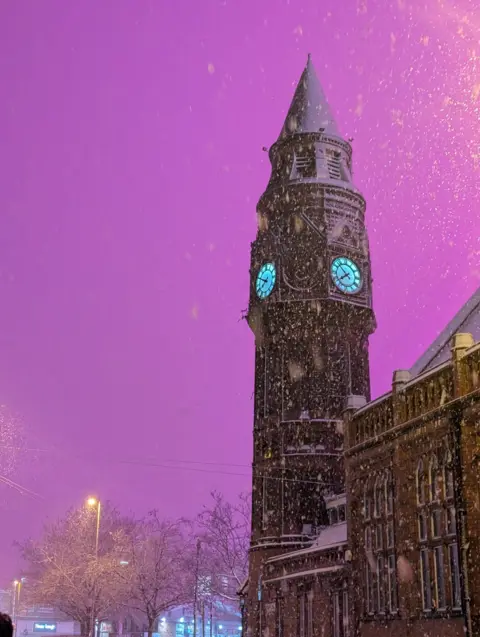 GLMCC Pink sky above a clock tower in Birmingham city centre. The face of the clock is lit and heavy snow can be seen falling around it. 