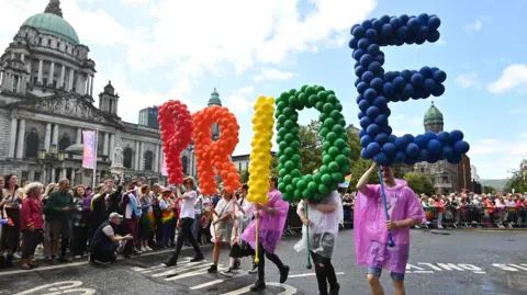 People carrying multicoloured balloons that spell out the word Pride. They are walking in front of Belfast City Hall.