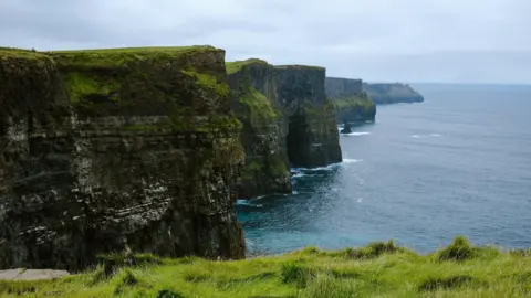 A view of the Cliffs of Moher on a cloudy day with a calm sea below.  The tall, rugged cliffs jut out at angles to the sea. 