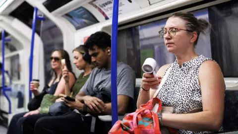 EPA A person uses an electric fan to cool down as commuters travel on the London Underground as the UK is set to experience its hottest June day on record during a heatwave in London, Britain, 24 June 2026. T