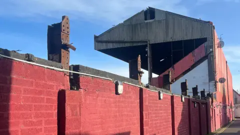 A red wall can runs from left to right where it meets a football stand. The stand is in a stae of disrepair