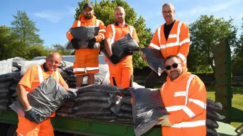 Five council workers in orange high vis outfits hold bags of black compost. Three stand on the back of a truck with the rest of the bags piled up. They are all smiling in the sunshine.