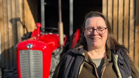 A women stands smiling in front of the tractor, she is wearing glasses as well as a body warmer and a fleece jacket. She has long dark hair
