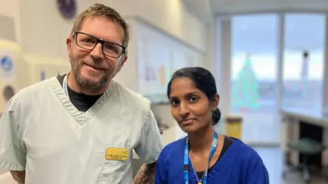 Two healthcare workers in a hospital setting, a man wearing a white overall and a woman wearing a blue overall, both smiling to camera