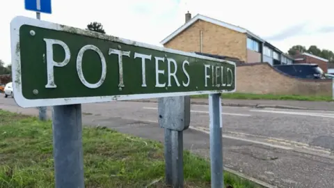 Andrew Turner/BBC A road sign bearing the name "Potters Field". A grass verge and pavement are in the foreground, with a terrace of 1970s houses opposite.