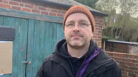 MICK LUNNEY/BBC Adam Dolling is pictured in a head and shoulders image. He is wearing an orangey-brown beanie and a black coat over a black hoodie. He is also wearing glasses and he has a fairly serious expression on his face. He is standing in what looks like a back yard. There is an outbuilding behind him and some trees in the background.