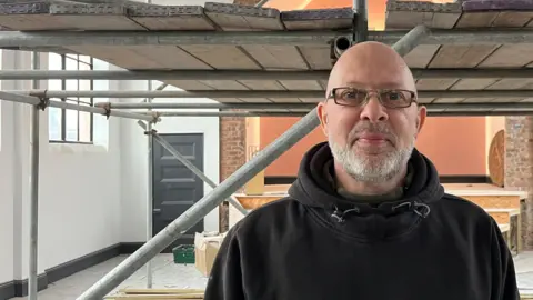 Mark Clarey with a grey beard wearing silver glasses. He is wearing a green fleece underneath a brown hoodie. He stands on the first-floor of the Amazing Graze building which he is helping to turn into a community centre and church. He is smiling.