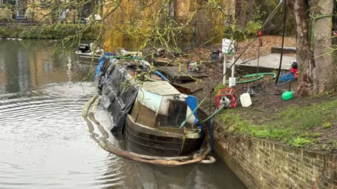 Narrowboat with green/brown, moss-covered at the top and a black hull, showing fire damage especially at the bow. There are black covers over the starboard side. It has been refloated and is resting on the surface of the water. There is a bund around the outside of the vessel. There is a smaller motor launch behind. There is a white sign to the right (which says "Strictly no mooring" and trees on the bank.