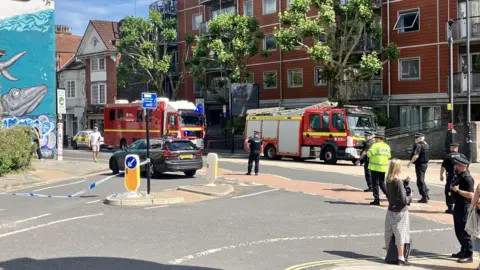 Two fire engines parked inside a cordon, with blue and white police tape securing a large section of the main road. 