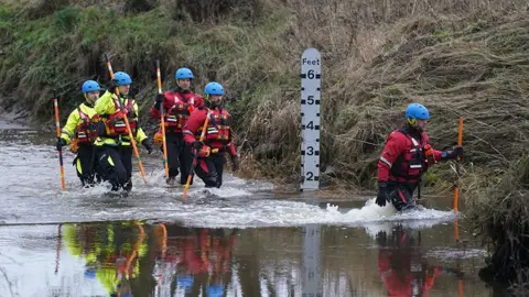 Five people dressed in life jackets, helmets and waterproof clothing are walking down a deep river carrying poles.