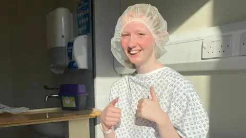 Enlli Thompson Enlli Thompson is photographed in hospital. She is smiling, looking at the camera with both thumbs up. She is wearing a hair net and a hospital gown.