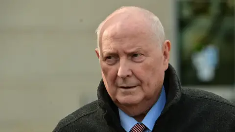 Pacemaker A man with balding white hair is looking off to the side of the camera. He is wearing a dark coloured coat, a blue shirt and a striped tie.
