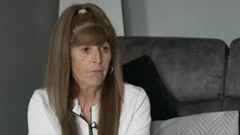 Lorraine Fraser has long, straight hair pulled back at the top with a hair tie, and the rest of the hair falls down past the shoulders. She is wearing a light-colored top with a zip at the front and a small microphone clipped near the neckline. A hoop earring is visible on one ear.
She is sitting on a large, dark upholstered chair with a soft texture.
