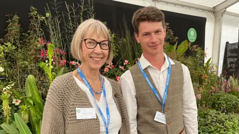 Clare Worden/BBC A woman and man stand in front of a display of plants. There are lots of different types of leaves, grasses and flowers behind them. She is wearing a light brown cardigan and white top. He is wearing a white shirt and a light brown waistcoat. 